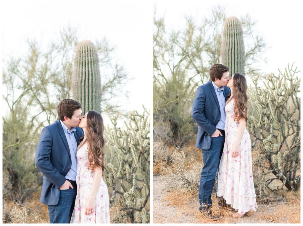 A man and a women with dark hair pose for their engagement photos holding hands and facing each other kissing in front of a cactus. He's in a blue suit jacket and she's in a light pink floral dress. 