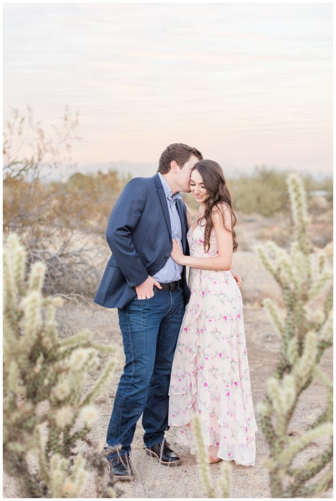 A man and a women with dark hair pose for their engagement pictures in  with cacti behind them. He's in a blue suit jacket and she's in a light pink floral dress. 