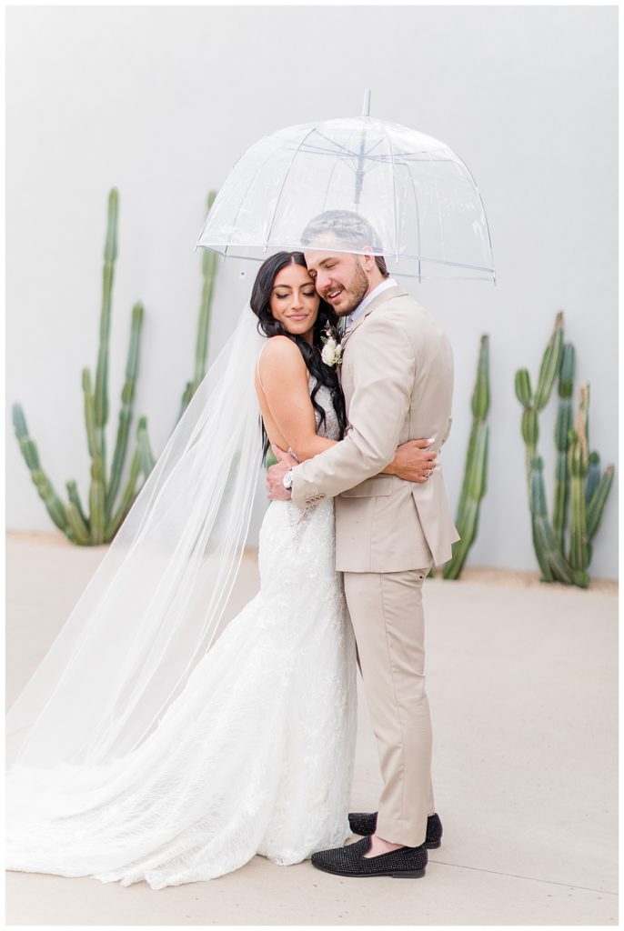 a man and woman in a wedding gown and light tan suit stand under an umbrella in an embrace in front of a gray wall and cacti 
