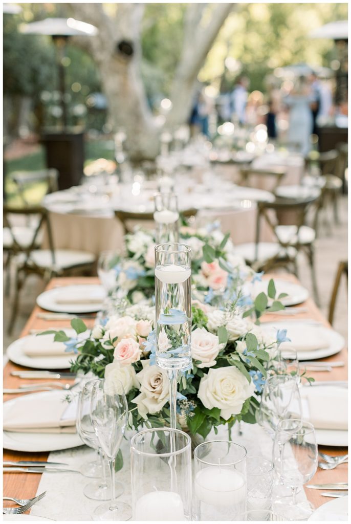 A table set up from an outdoor reception with light pink and white flowers, greenery, plate settings, and white floating candles. 