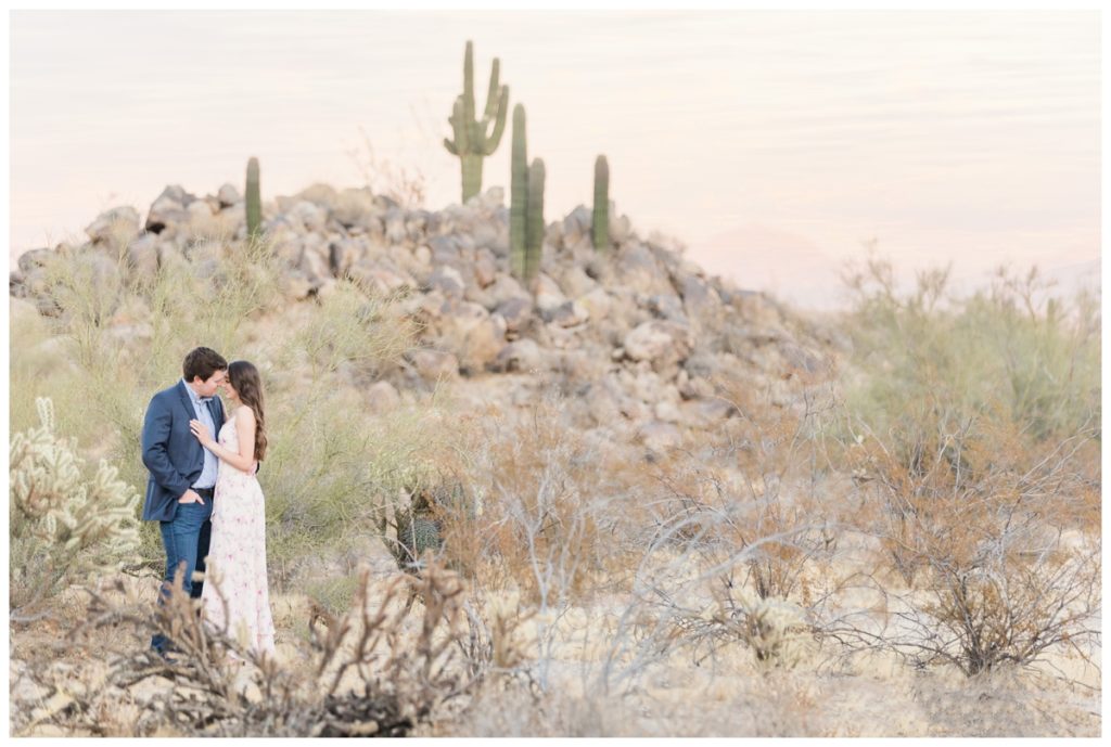 A man and a women with dark hair pose for their engagement photos in the desert with cacti behind them. He's in a blue suit jacket and she's in a light pink floral dress. 