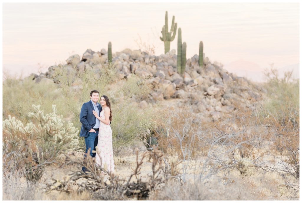 A man and a women with dark hair pose for their engagement photos in the desert with cacti behind them. He's in a blue suit jacket and she's in a light pink floral dress. 